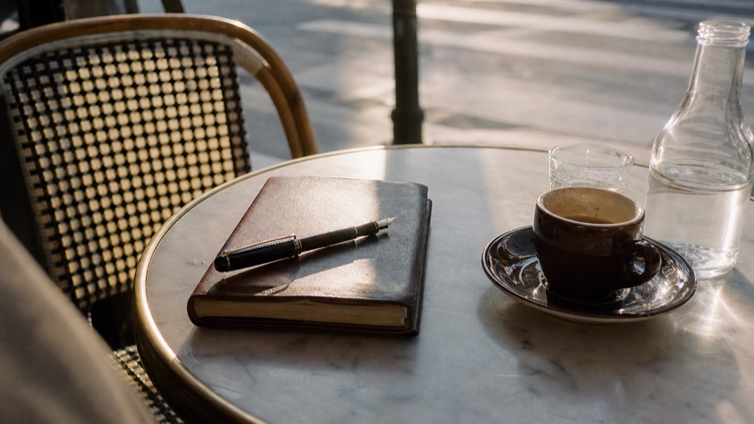 Morning light on a closed leather notebook and pen beside an espresso on a marble Paris cafe table